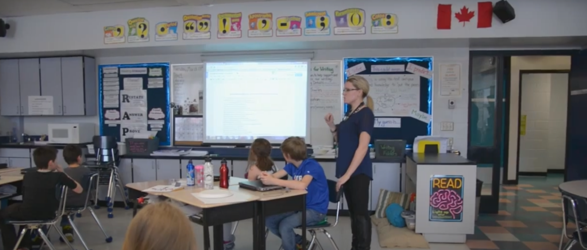Classroom of adolescent students with an adult female teacher standing in the centre of the room with arm raised in conversation. Behind the teacher is a classroom screen, notation boards, and posters.