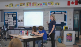 Classroom of adolescent students with an adult female teacher standing in the centre of the room with arm raised in conversation. Behind the teacher is a classroom screen, notation boards, and posters.
