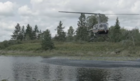 Image shows a helicopter flying over a shallow lake with green forested landscape in the background