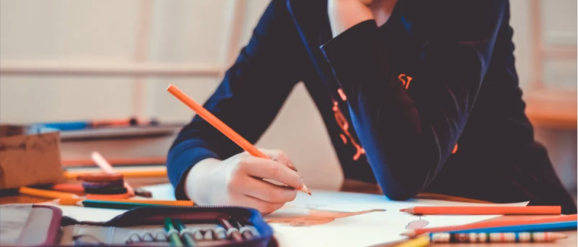 Child seated at desk in front profile holding a pencil to paper with assorted stationary around them.