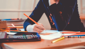 Child seated at desk in front profile holding a pencil to paper with assorted stationary around them.