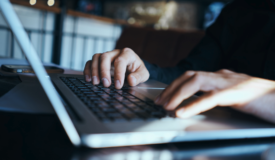 Hands typing on white laptop in left profile with Iphone in background.
